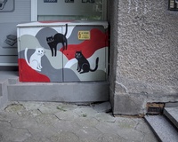 A street electrical box decorated with a picture of three cats, two black and one white, on an abstract wave-like background